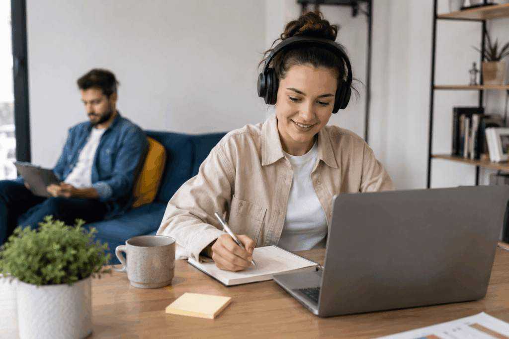 A imagem mostra uma profissional trabalhando em casa com notebook, fones de ouvido e materiais de organização. Ao fundo, outra pessoa também realiza uma atividade profissional em um ambiente confortável. A cena transmite a ideia de flexibilidade, autonomia e integração entre vida profissional e rotina doméstica.