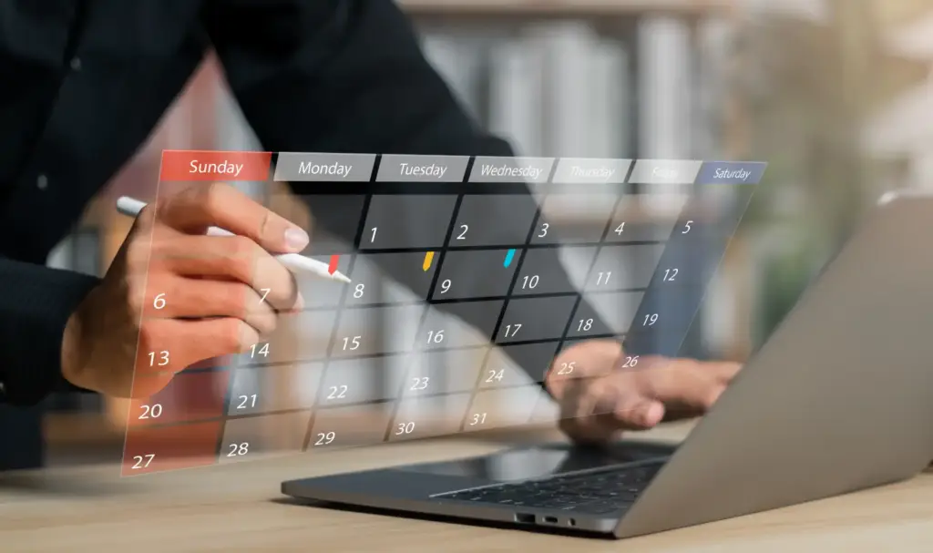 Pessoa utiliza um tablet transparente para marcar compromissos no calendário digital, enquanto trabalha em um laptop na mesa.