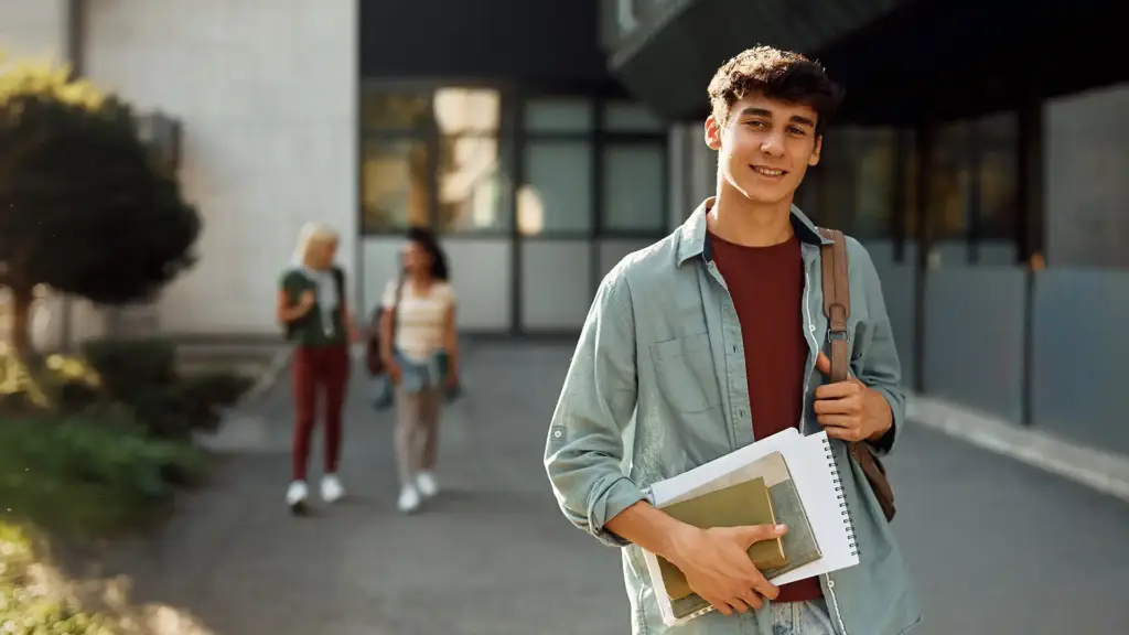 Estudante sorridente segurando livros e caderno, caminhando ao lado de colegas na universidade, em um ambiente externo com prédio moderno ao fundo.