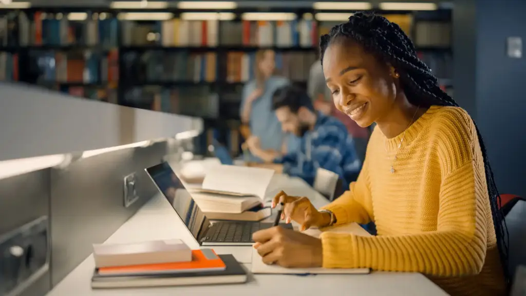 Jovem estudante sorridente estudando com livros e laptop em uma biblioteca, focada na palavra-chave 'estudo e aprendizado' em ambiente acadêmico.