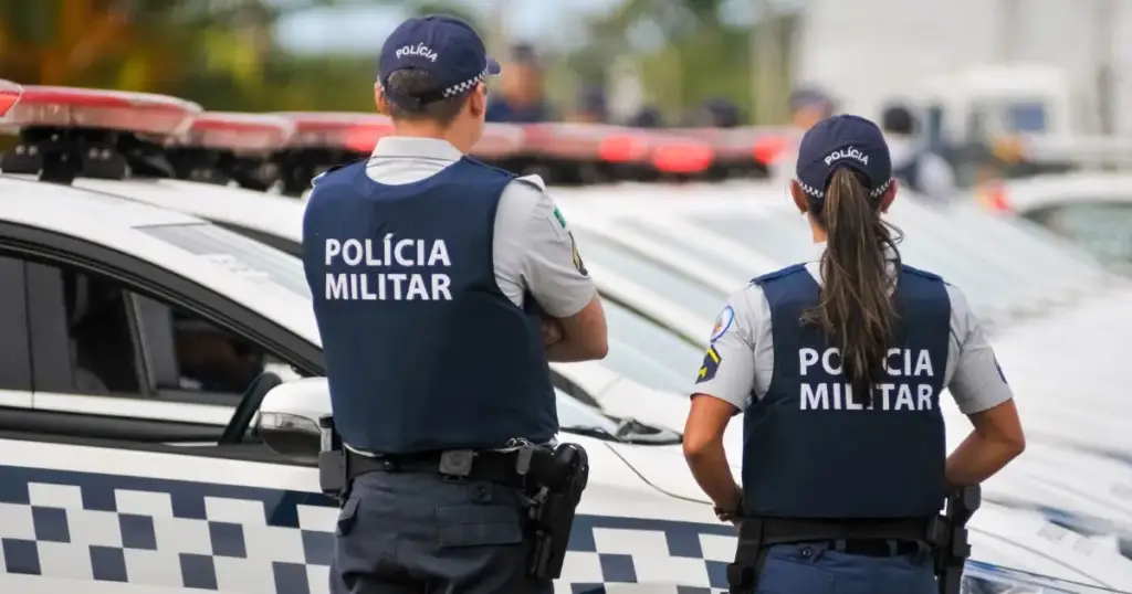 Dois policiais militares de costas ao lado de uma viatura policial